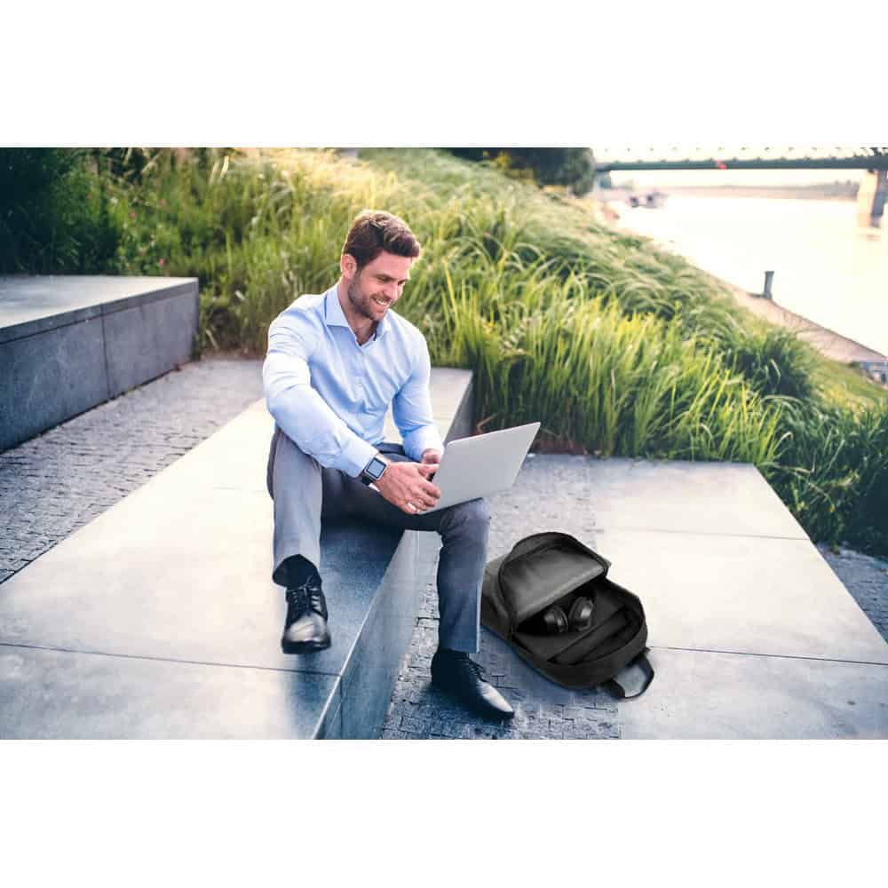 Man med laptop och hörlurar utomhus, ryggsäck bredvid på stenbänk.